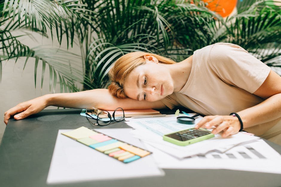A tired woman calculating her taxes at a desk covered with papers and a smartphone.