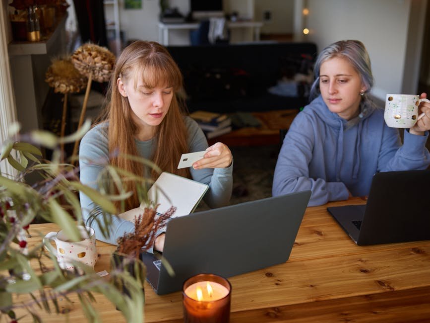 Two women using laptops for online shopping at home, holding credit cards and coffee mugs.