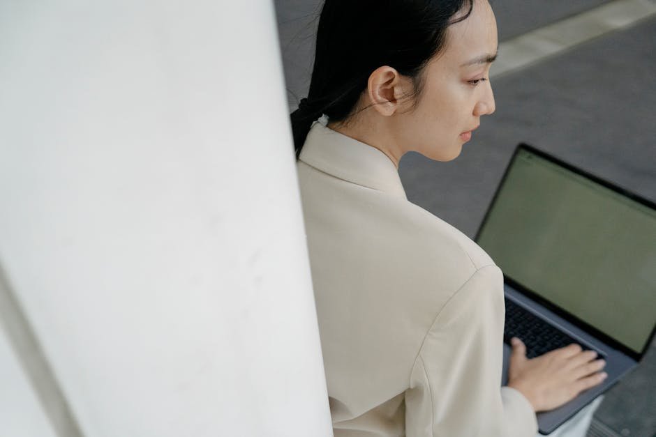 From above of crop concentrated young Asian female freelancer with long dark hair in elegant outfit working remotely on laptop while sitting on bench on street