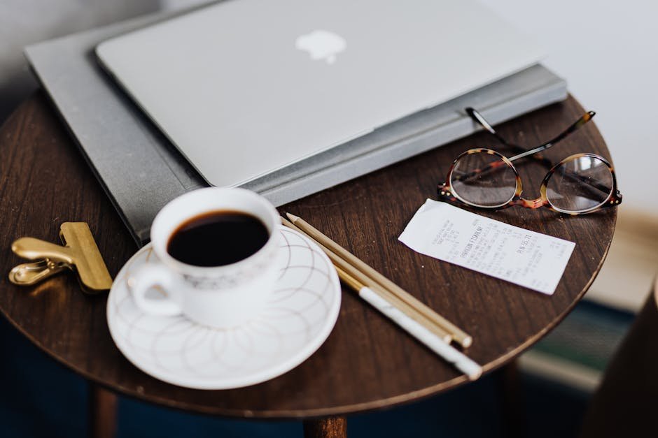 A close-up of a wooden table with a laptop, coffee, eyeglasses, and stationery, creating a cozy workspace vibe.