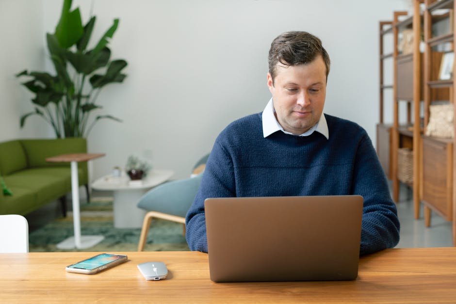 Caucasian man using a laptop in a modern living room setting with plants and shelves.
