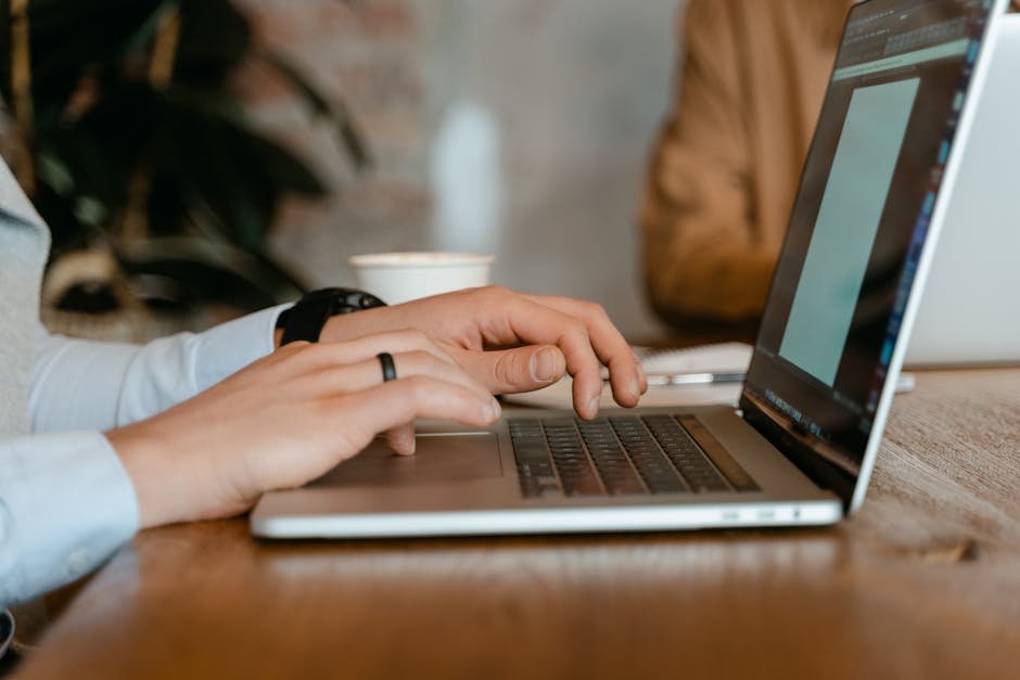 A person typing on a laptop at a wooden table in a modern office setting.