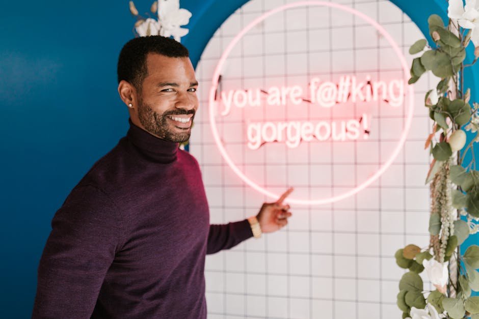 Portrait of a smiling man in a hair salon pointing at a neon sign with positive message.