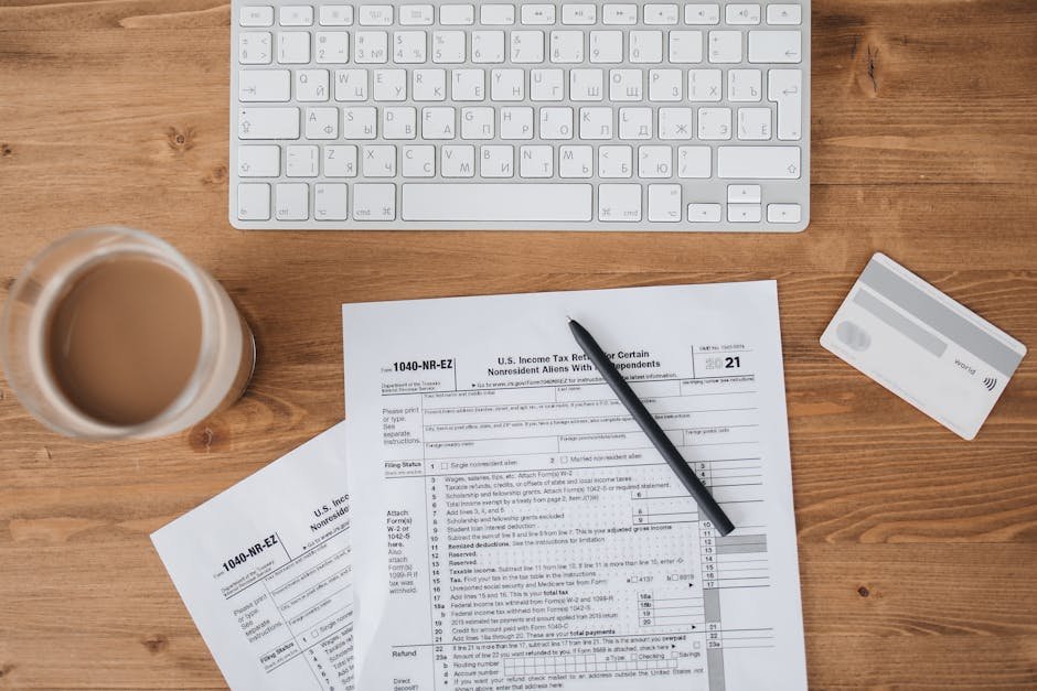 Top view of tax forms, keyboard, pen, and coffee on a wooden desk.