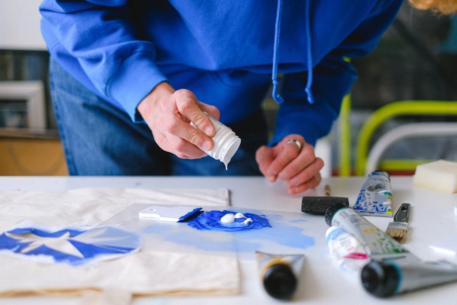 Unrecognizable male master with small jar adding white paint into blue pigment while working at table with stencil printing in studio on blurred background