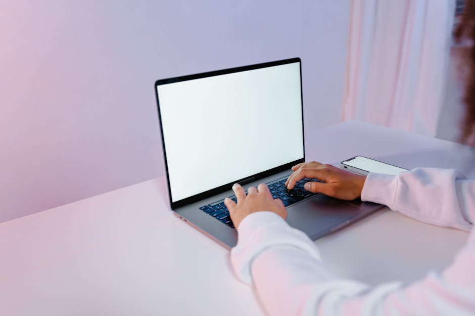Hands typing on a laptop with a blank screen on a white desk, smartphone nearby.