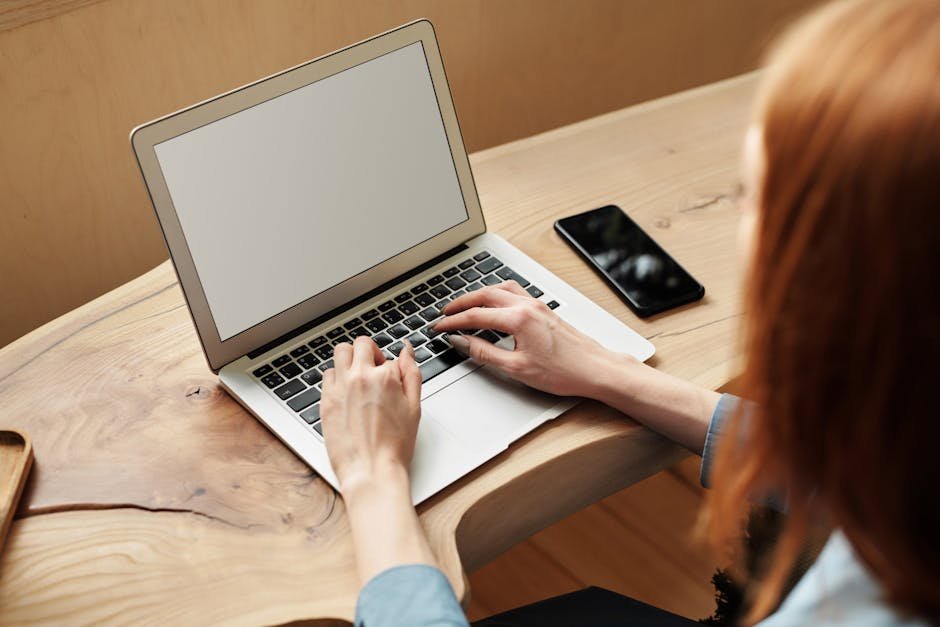 A woman working on a laptop at a wooden desk, indoors with natural light.