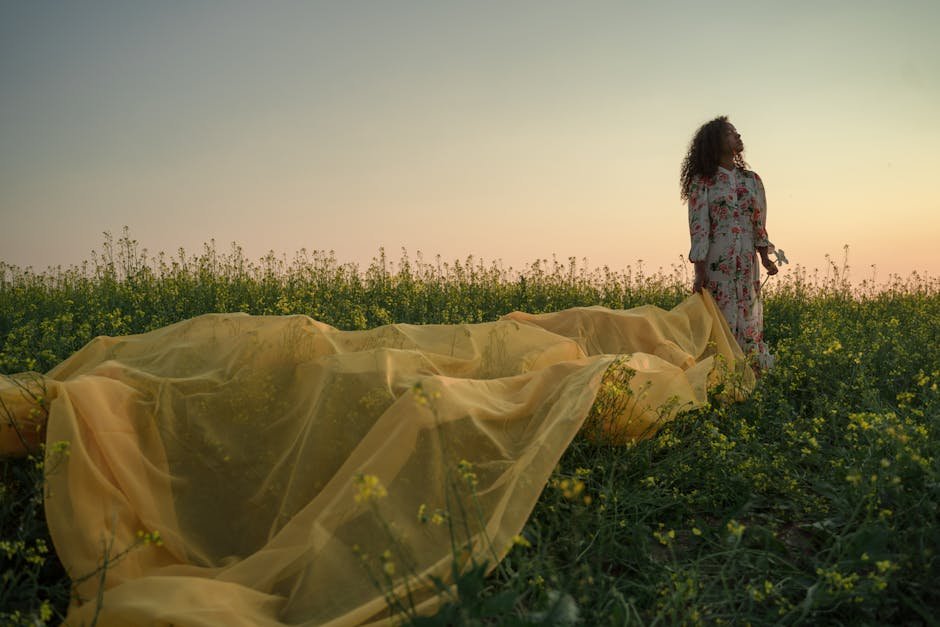 A woman stands in a field with yellow fabric and flowers during sunset, conveying tranquility.