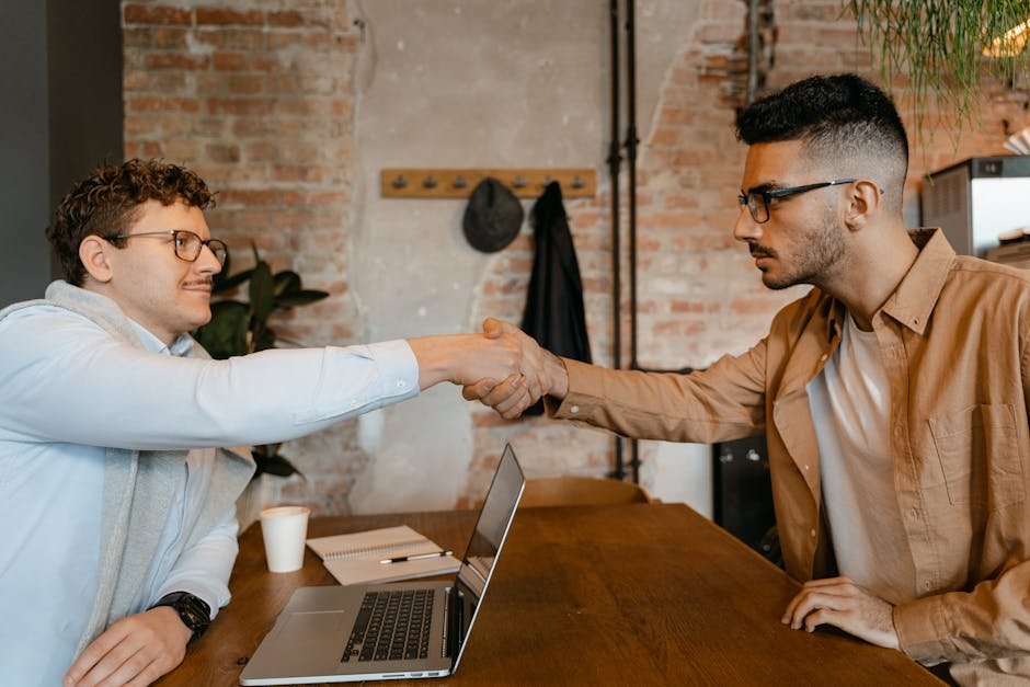 Businessmen shaking hands over a laptop at a wooden table in a modern office setting.